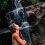 Side view of smiling traveling nude female covering breast and standing in calm water of pond near waterfall in Fervenza De Casarinos natural park in Spain location traveler