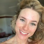 a woman with blonde hair smiles while she stands in front of her bed Porn Pic
