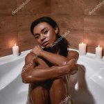 Young beautiful and sexy mixed race nude woman sitting in the bath with candles at home looking at camera Stock Photo LanaStock 449393654
